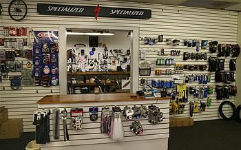 Image of a bicycle shop interior featuring a service counter made of wood, with various bicycle accessories and tools displayed. The walls are lined with organized shelves showcasing a range of bike parts and equipment, branded as "Specialized." The atmosphere is well-lit and presents a focus on cycling merchandise.