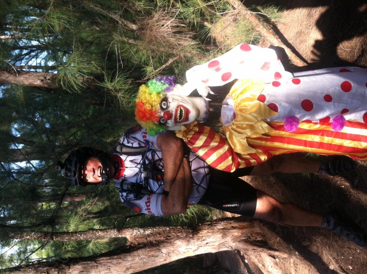 A person wearing a cycling outfit poses with a clown in a colorful costume and wig, both standing in a wooded area. The clown features a bright makeup design with a wide grin, and they are surrounded by trees and natural surroundings. Oleta River State Park mountain bike trail.