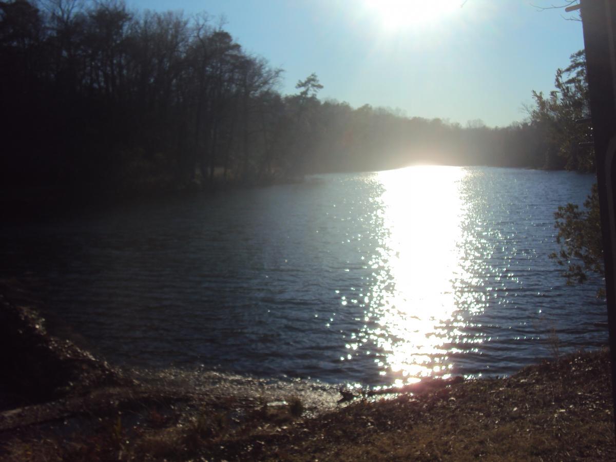 A tranquil view of a lake shimmering under the sunlight, surrounded by trees in the background. The sun is positioned high in the sky, casting a bright reflection on the water's surface. The scene conveys a peaceful, natural setting. Yorktown Historical Trail mountain bike trail.