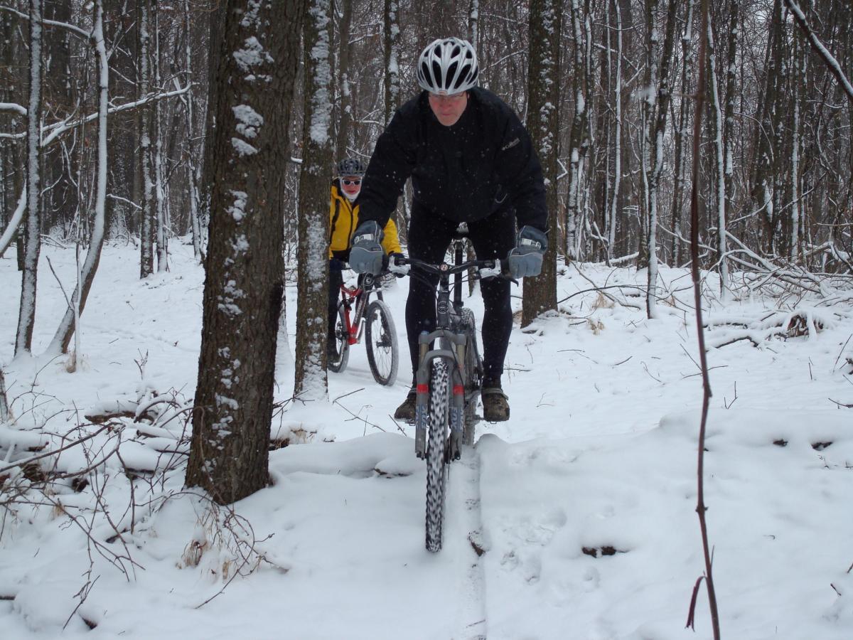A mountain biker navigates a snowy forest trail, focused on maintaining balance as he rides over a narrow path. In the background, another cyclist in yellow gear follows closely, surrounded by trees and a blanket of snow covering the ground. Hartwood Acres mountain bike trail.