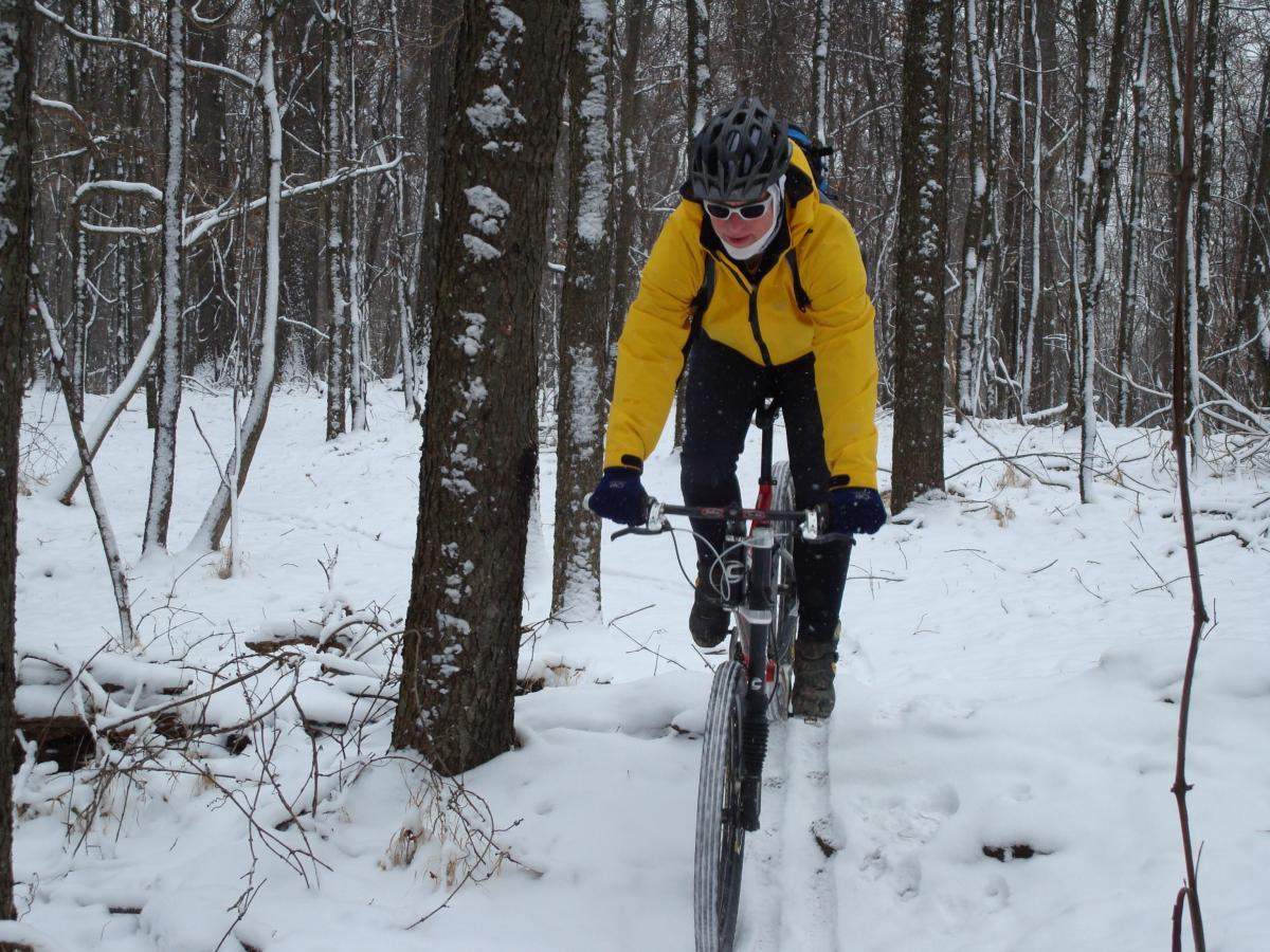 A person riding a mountain bike through a snowy forested area, wearing a yellow jacket, black pants, gloves, and a helmet. Snow is covering the ground and trees, creating a winter landscape. The cyclist is focused on the path ahead as they navigate through the snow. Hartwood Acres mountain bike trail.