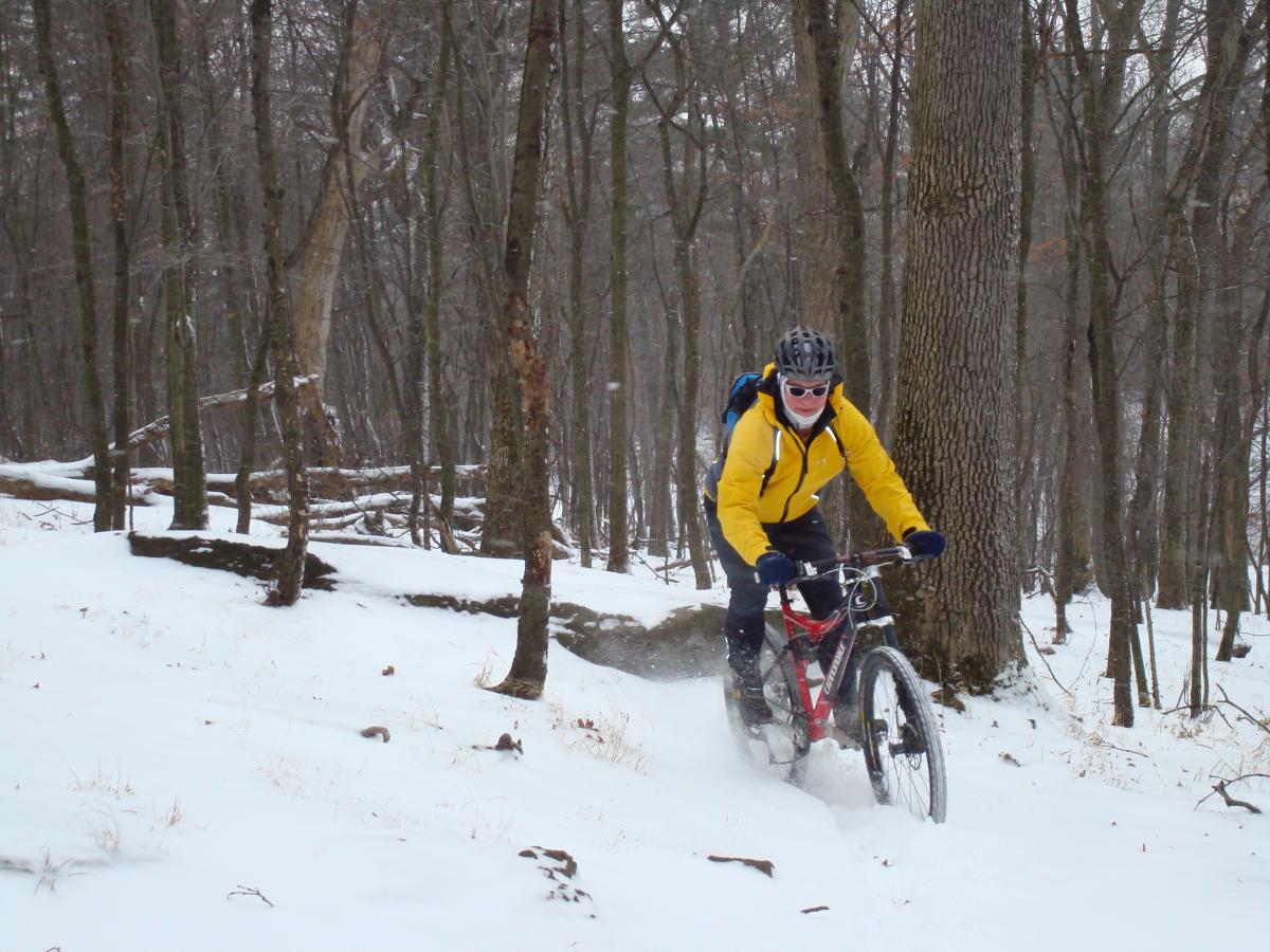 A person wearing a bright yellow jacket and sunglasses is riding a mountain bike through a snowy forest trail. Surrounding trees are bare, indicating winter, and the ground is covered in a layer of snow. The rider appears to be navigating a slight downhill slope, with snow spraying up from the bike’s tires. Hartwood Acres mountain bike trail.