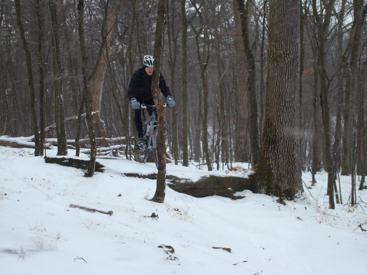 A cyclist navigating through a snowy forest trail, surrounded by trees and winter scenery. Hartwood Acres mountain bike trail.