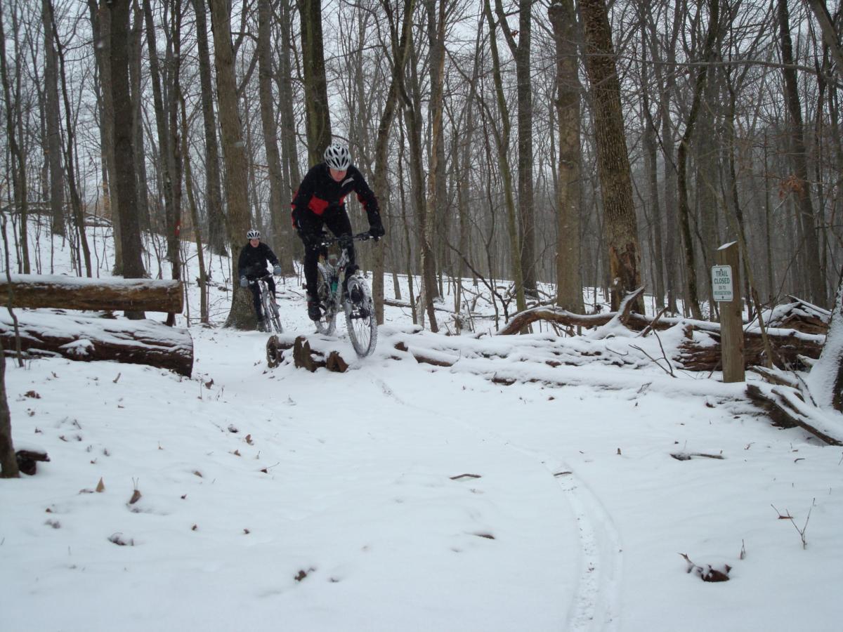 Two mountain bikers navigate a snow-covered trail in a wooded area during winter. One biker is in the foreground, leaping over a log, while the other follows closely behind. Snow blankets the ground and trees, creating a serene but challenging winter landscape. A "Trail Closed" sign is visible on the right side of the image. Hartwood Acres mountain bike trail.