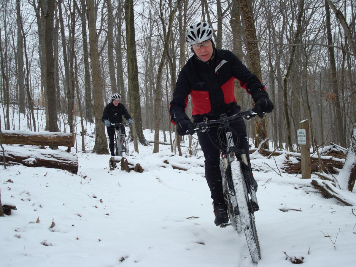 Two mountain bikers riding through a snowy forest trail. One rider is in the foreground, wearing a helmet and a black jacket with red accents, focused on navigating the snowy path. The second rider is in the background, slightly behind, also wearing winter biking gear. Snow covers the ground, and trees surround the trail, with a sign indicating a closed trail visible nearby. Hartwood Acres mountain bike trail.
