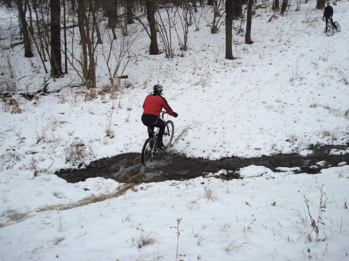 A person in a red jacket rides a mountain bike through a shallow stream, surrounded by snowy terrain and sparse trees. In the background, another cyclist can be seen approaching the same area. Hartwood Acres mountain bike trail.