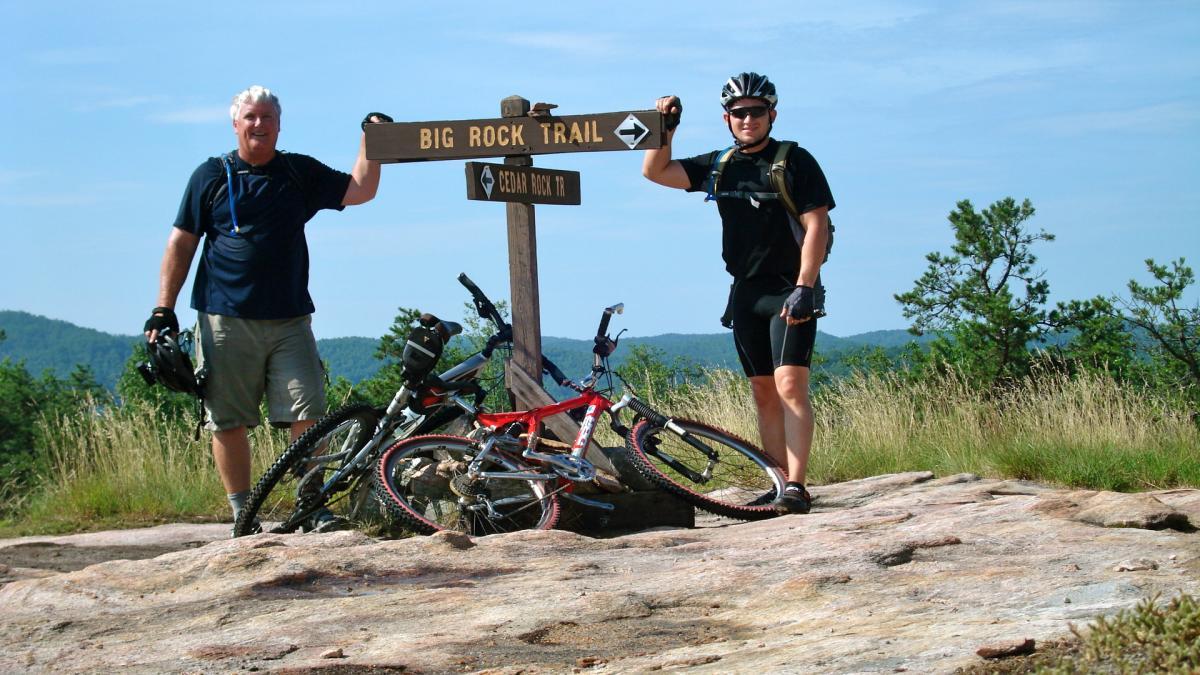 Two mountain bikers stand next to a wooden signpost marking the Big Rock Trail, with their bikes parked nearby. The scene is set in a natural outdoor environment, featuring rolling hills and greenery in the background under a clear blue sky. Cedar Rock Trail #16 mountain bike trail.