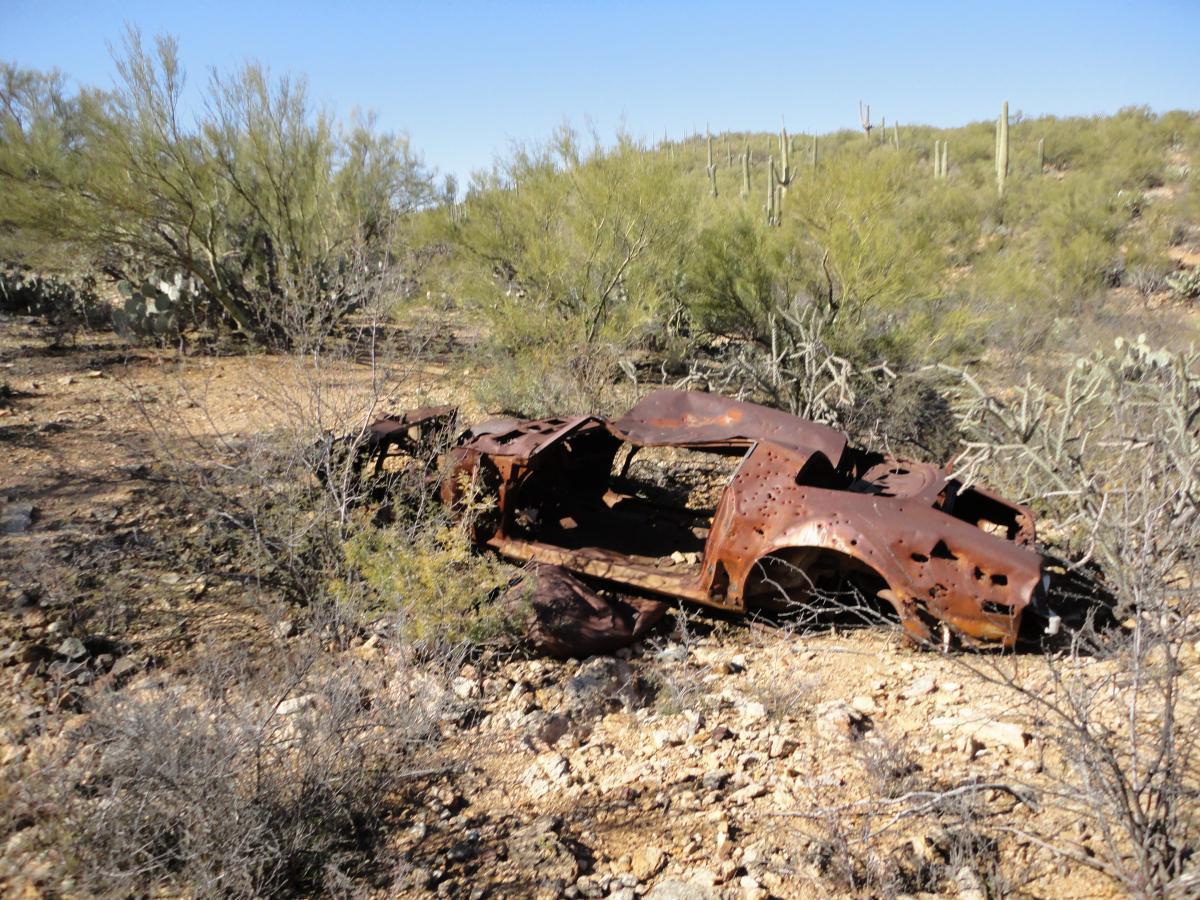 Alt text: Rusted remains of an old car are partially buried in a desert landscape, surrounded by sparse vegetation and small bushes, under a clear blue sky. Robles Pass mountain bike trail.