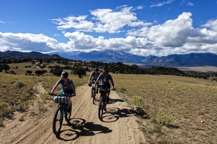 Four mountain bikers riding on a dirt trail in a scenic landscape with rolling hills and mountains in the background under a blue sky with scattered clouds.