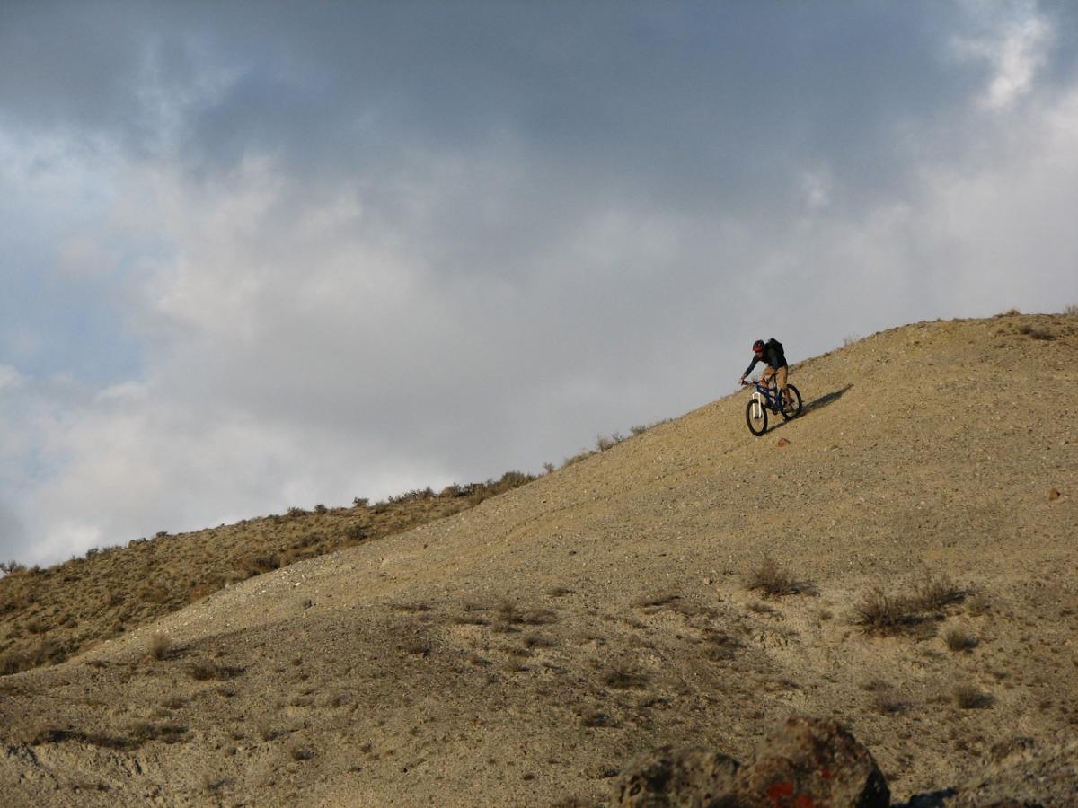 A mountain biker traversing a rocky hillside under a partly cloudy sky. The terrain is dry and rugged, with sparse vegetation visible on the slopes. The biker leans forward, navigating the incline. Discovery Hill Trails mountain bike trail.