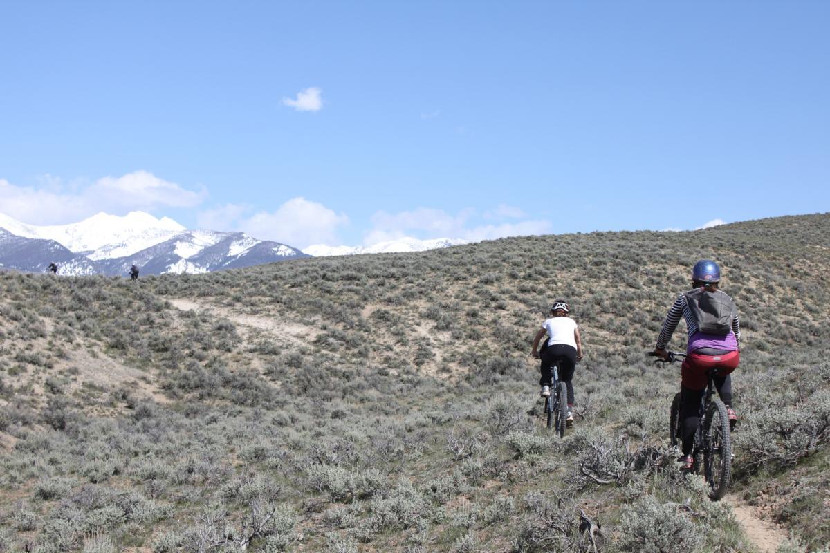 Two mountain bikers riding on a dirt trail through sagebrush-covered hills, with snow-capped mountains in the background under a blue sky with a few clouds. Discovery Hill Trails mountain bike trail.