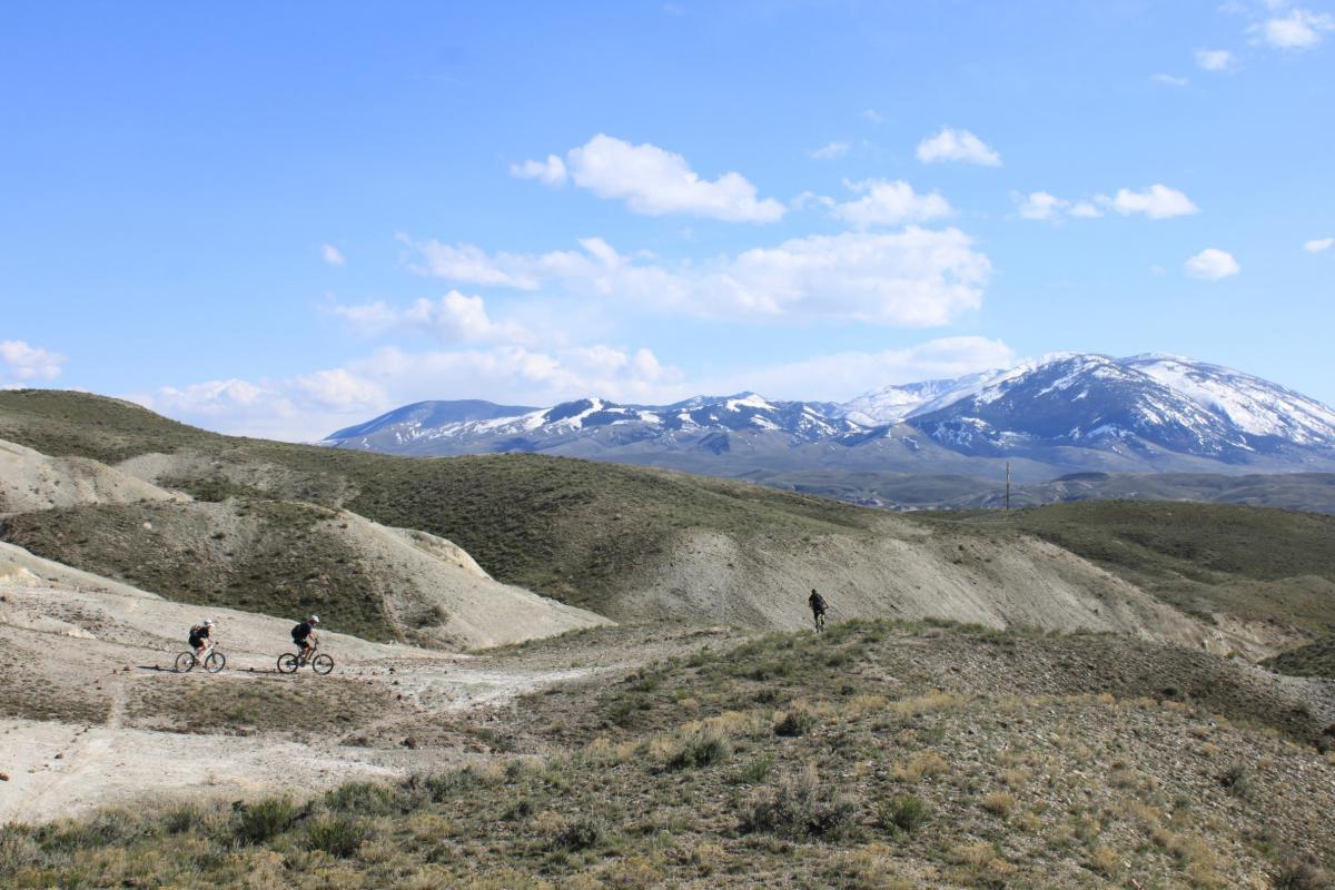 Two mountain bikers ride along a dirt path in a hilly, grassy landscape, with snow-capped mountains in the background under a blue sky with scattered clouds. Discovery Hill Trails mountain bike trail.