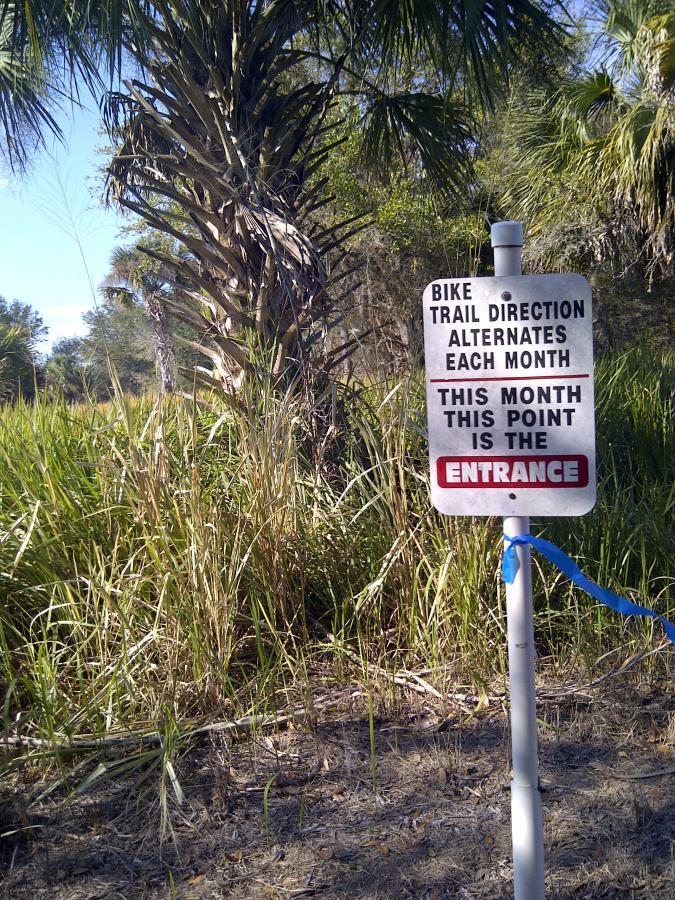 A sign indicating the entrance to a bike trail, stating "Bike trail direction alternates each month. This month this point is the ENTRANCE," surrounded by dense greenery and palm trees. Caloosahatchee Regional Park mountain bike trail.
