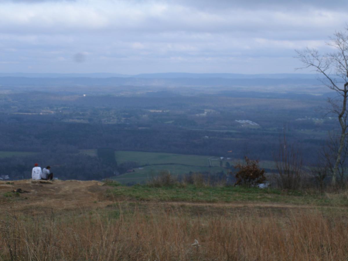 Two people sitting on a hillside, overlooking a vast landscape of rolling hills and valleys under a cloudy sky. The foreground features tall grass and the distant view includes patches of trees and open fields. Pigeon Mountain mountain bike trail.