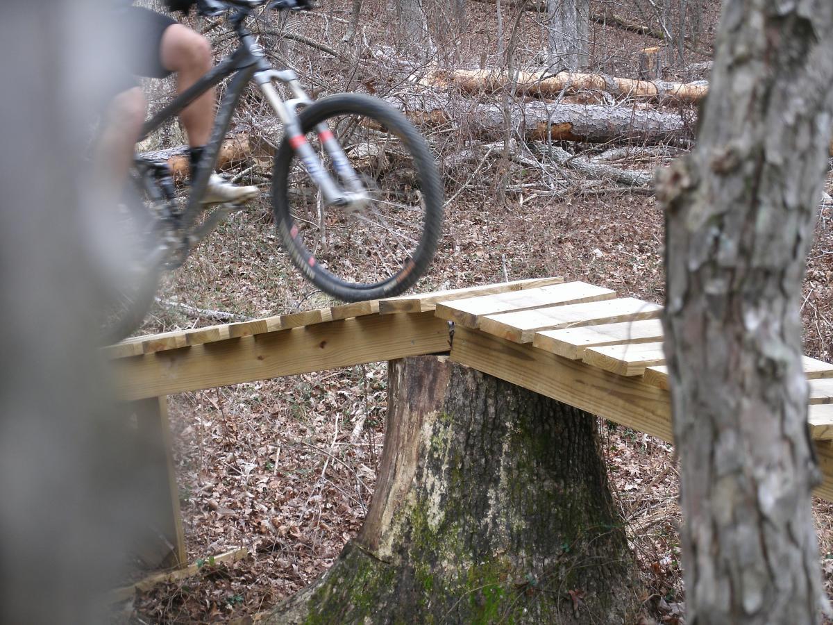 A mountain biker navigating a wooden bridge over a wooded area, with trees and fallen branches in the background. The cyclist is mid-ride, captured in action. Enterprise South mountain bike trail.