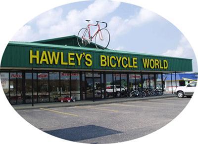 Exterior view of Hawley's Bicycle World, a bike shop featuring a large green building with the store name prominently displayed in yellow letters. A pink bicycle is mounted on the roof, and various bicycles are visible in the display windows. The surrounding area has a paved parking lot.