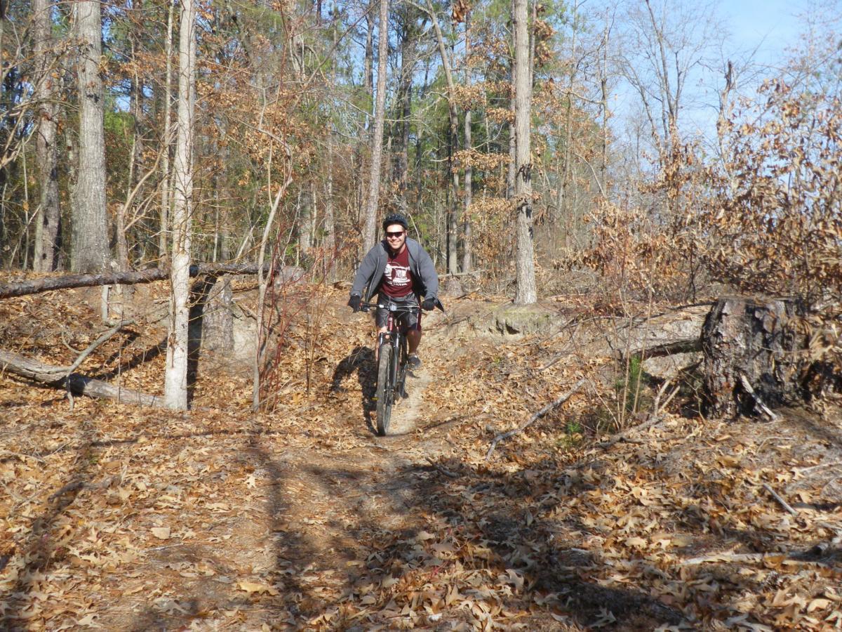A person riding a mountain bike on a dirt trail through a wooded area, surrounded by trees with sparse foliage and fallen leaves. Lake Rim Park Trail mountain bike trail.