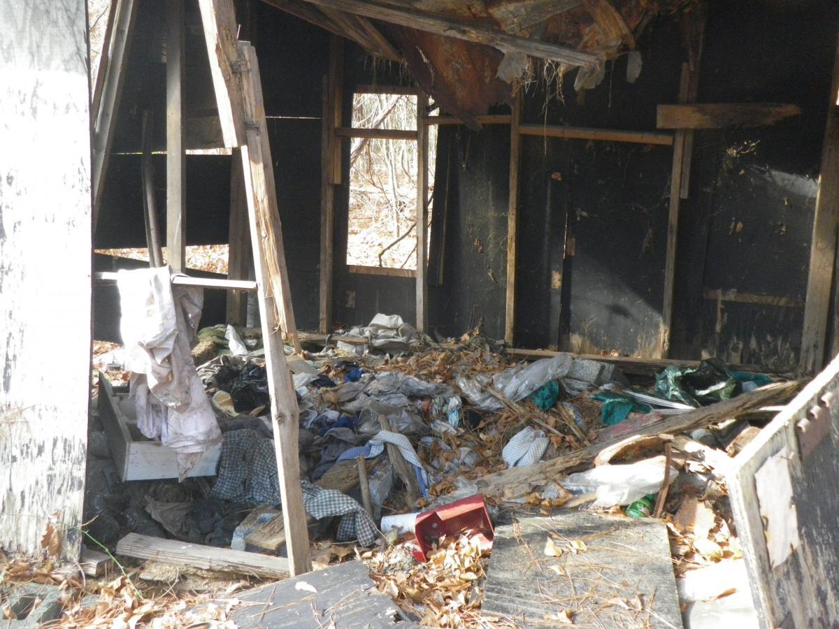 Alt text: An abandoned, dilapidated interior of a small structure filled with clutter, including piles of discarded clothing and debris, with sunlight filtering through the gaps in the walls. Lake Rim Park Trail mountain bike trail.