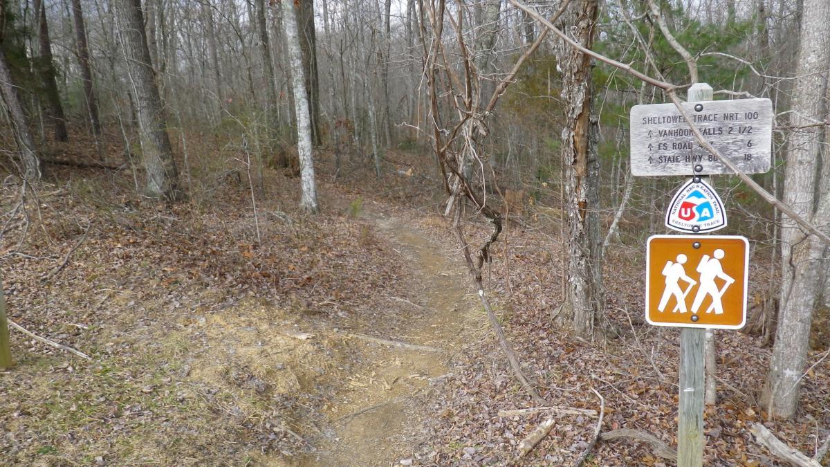 A dirt hiking path leading into a wooded area, marked by a trail sign indicating directions to nearby locations, including Vanhook Falls and State Highway 80. A hiker symbol sign is visible next to the trailhead. The scene shows bare trees and scattered leaves on the ground, suggesting a late autumn or early spring atmosphere. Cane Creek (sheltowee Trace Trail) mountain bike trail.