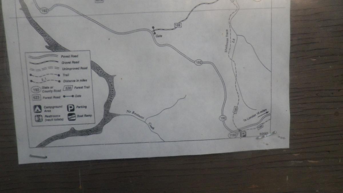 A printed map displaying roads, trails, and points of interest, including a campground area, parking, and a boat ramp. The map features various road types, such as paved, gravel, and unpaved roads, with distance markers indicated in miles. Key locations and a gate are also marked, alongside a reference guide for interpreting the map symbols. Cane Creek (sheltowee Trace Trail) mountain bike trail.
