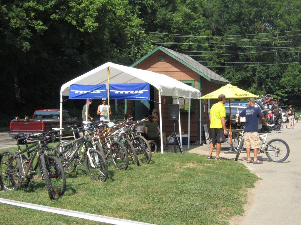 A summer outdoor scene featuring a tent set up with the brand name "Titus." In front of the tent, several mountain bikes are lined up. People are gathered near the tent, some engaged in conversation, while others are preparing to ride their bikes. A small building with green accents is visible in the background, surrounded by trees.
