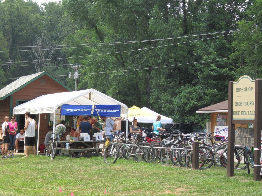 A busy bike shop scene featuring a group of people gathered under a canopy tent, with various bicycles parked nearby. The shop offers bike rentals and tours, and there are signs indicating the services available. The setting is surrounded by lush greenery, creating a vibrant outdoor atmosphere.