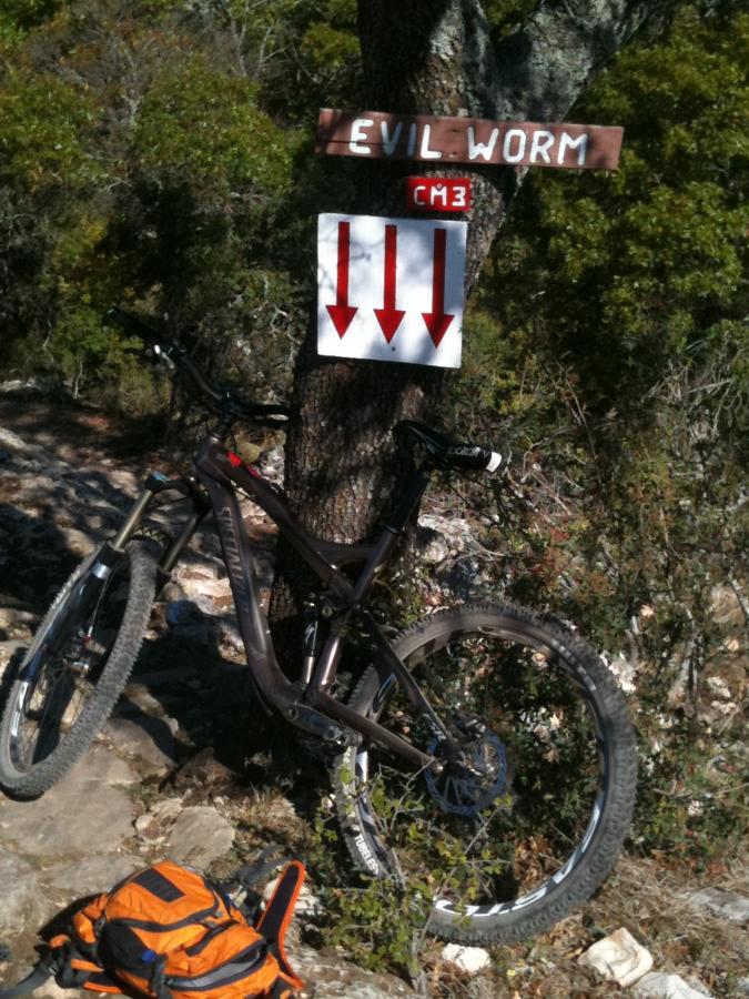 A mountain bike leaning against a tree with a wooden sign reading "Evil Worm" and directional arrows pointing downward, surrounded by greenery and rocky terrain. An orange backpack is visible on the ground in the foreground. Flat Rock Ranch mountain bike trail.