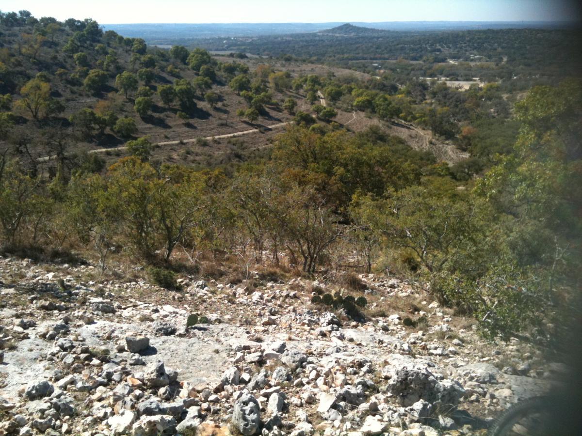 A scenic view from a rocky hillside, featuring a diverse landscape of rolling hills, scattered trees, and a dirt road winding through the terrain. The image captures a sunny day, highlighting the natural beauty of the area with clear skies and distant mountains visible in the background. Flat Rock Ranch mountain bike trail.