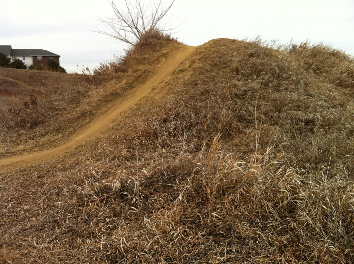 A dirt mound with a gradual incline, surrounded by dry grass, set against a cloudy sky. In the background, a residential building is partially visible. The scene suggests a natural landscape, potentially used for recreational activities such as biking or hiking. Tranquility mountain bike trail.