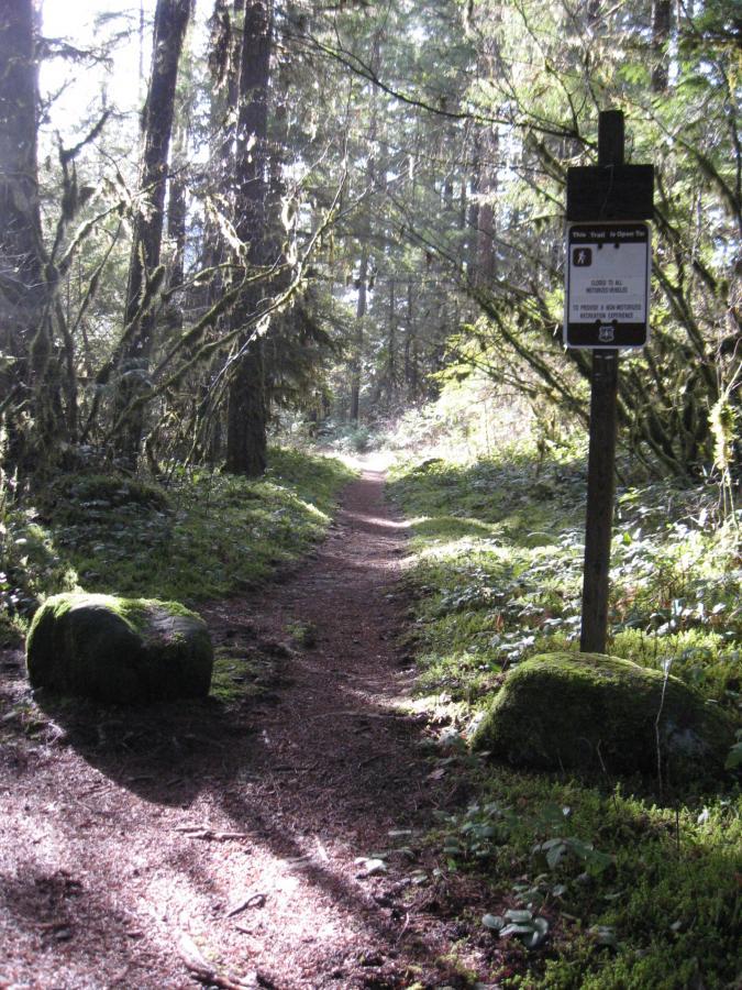 A narrow, winding dirt path leads through a lush, green forest. Tall trees line the trail, their branches partially obscuring the sunlight. On the right side of the path, a wooden sign provides information about the area, while moss-covered rocks are visible on either side of the trail. The scene conveys a tranquil and inviting atmosphere in nature. Salmon Creek Trail mountain bike trail.
