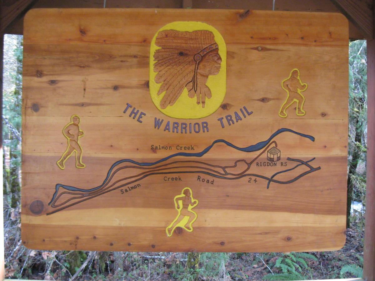 A wooden sign depicting "The Warrior Trail." The sign features an illustration of a Native American headdress at the top center. Below the headdress, the trail and surrounding features are outlined with blue lines, showing the route along Salmon Creek Road. Yellow figures are depicted on either side, representing runners. The sign includes the text "THE WARRIOR TRAIL" and names the locations "Salmon Creek" and "Rigdon RS." The sign has a rustic appearance, situated in a natural setting with trees in the background. Salmon Creek Trail mountain bike trail.