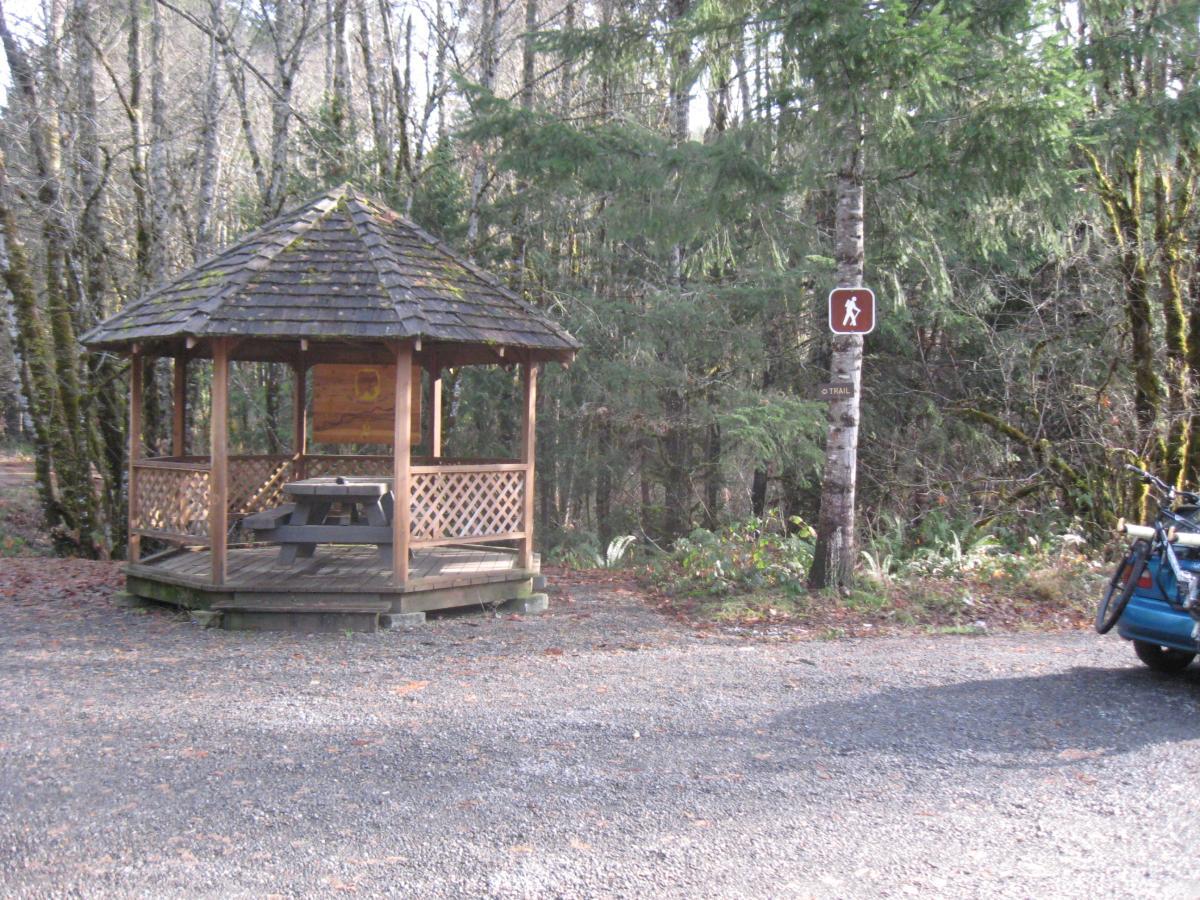 A wooden gazebo with a shingled roof, featuring a picnic table inside, set against a backdrop of trees. A trail sign is visible nearby, indicating a hiking path. The ground is gravel, and fallen leaves are scattered around. Salmon Creek Trail mountain bike trail.
