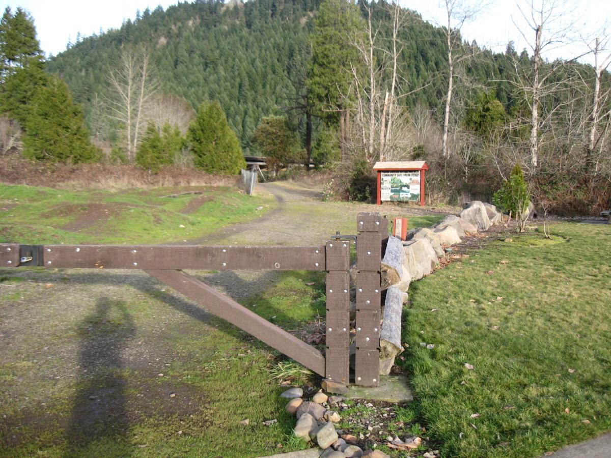 A wooden gate stands at the entrance to a gravel path leading into a green, hilly landscape. In the background, there is a sign indicating the location, surrounded by trees and shrubs. The grass is lush, and a rocky border delineates the area. This peaceful outdoor scene suggests a recreational or natural area. Alpine Trail mountain bike trail.