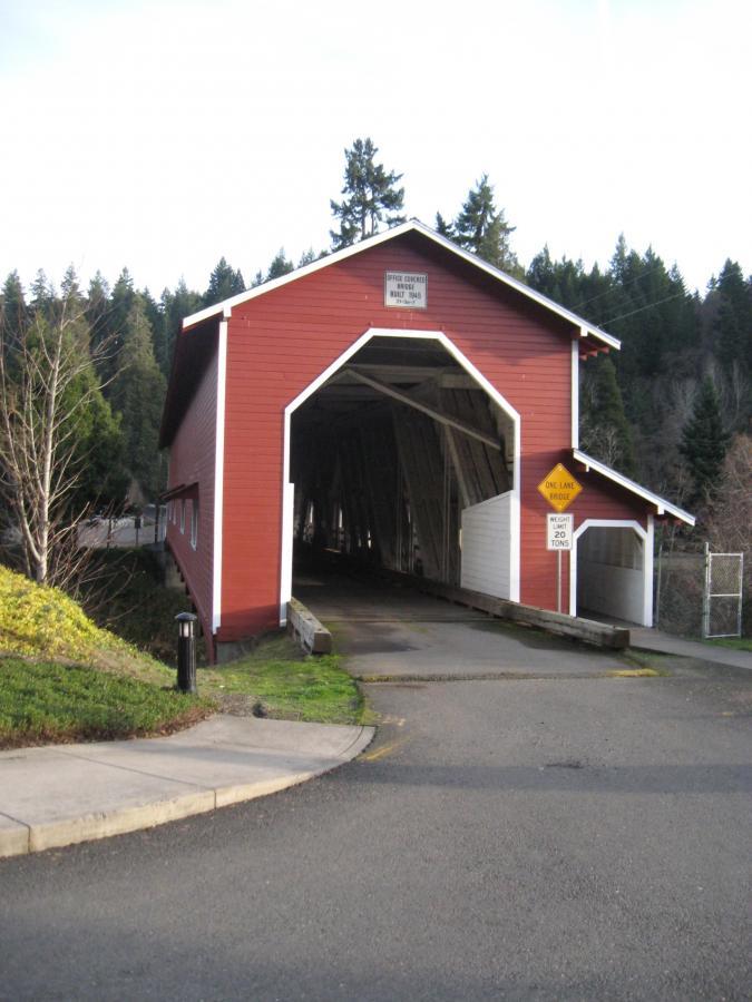 A red covered bridge spanning a road, with a sign indicating it's a one-lane bridge. The surrounding landscape features trees in the background and a paved path leading to the bridge. Alpine Trail mountain bike trail.