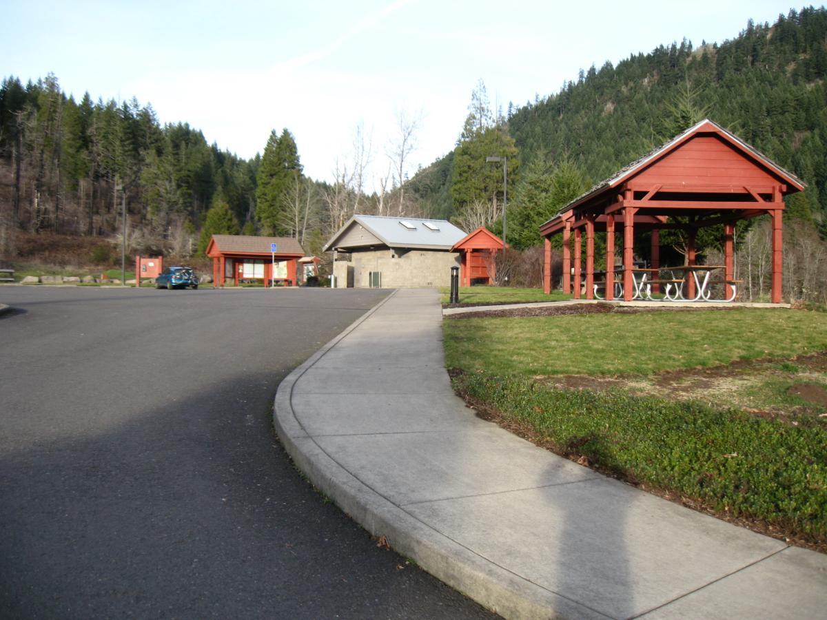 Alt text: "A park area featuring a paved walkway leading to red-roofed buildings, including a restroom facility and a covered picnic area. Lush green trees line the background, indicating a forested environment." Alpine Trail mountain bike trail.