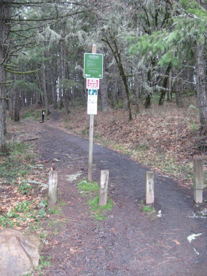 A dirt path leading through a wooded area, featuring a signpost with multiple informational signs. The path is flanked by wooden posts and surrounded by trees with a carpet of fallen leaves. The atmosphere appears to be calm and natural, suggesting a hiking or walking trail. Ridgeline Trail mountain bike trail.