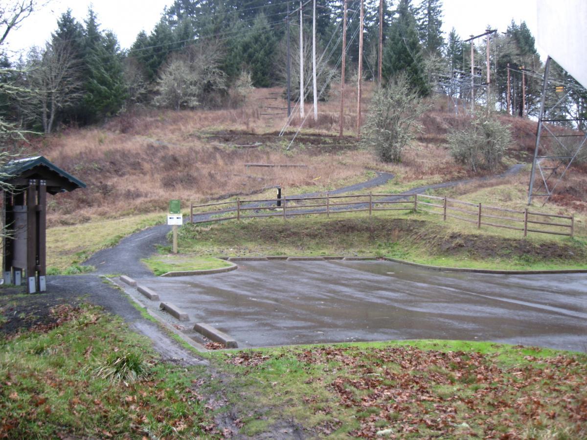 A scenic view of a partially wet parking area leading to a hiking trail, surrounded by grassy land and sparse trees. A wooden fence lines the path that extends up a hill, while a signpost is visible near the trailhead. The landscape suggests a rural outdoor setting on a cloudy day. Ridgeline Trail mountain bike trail.