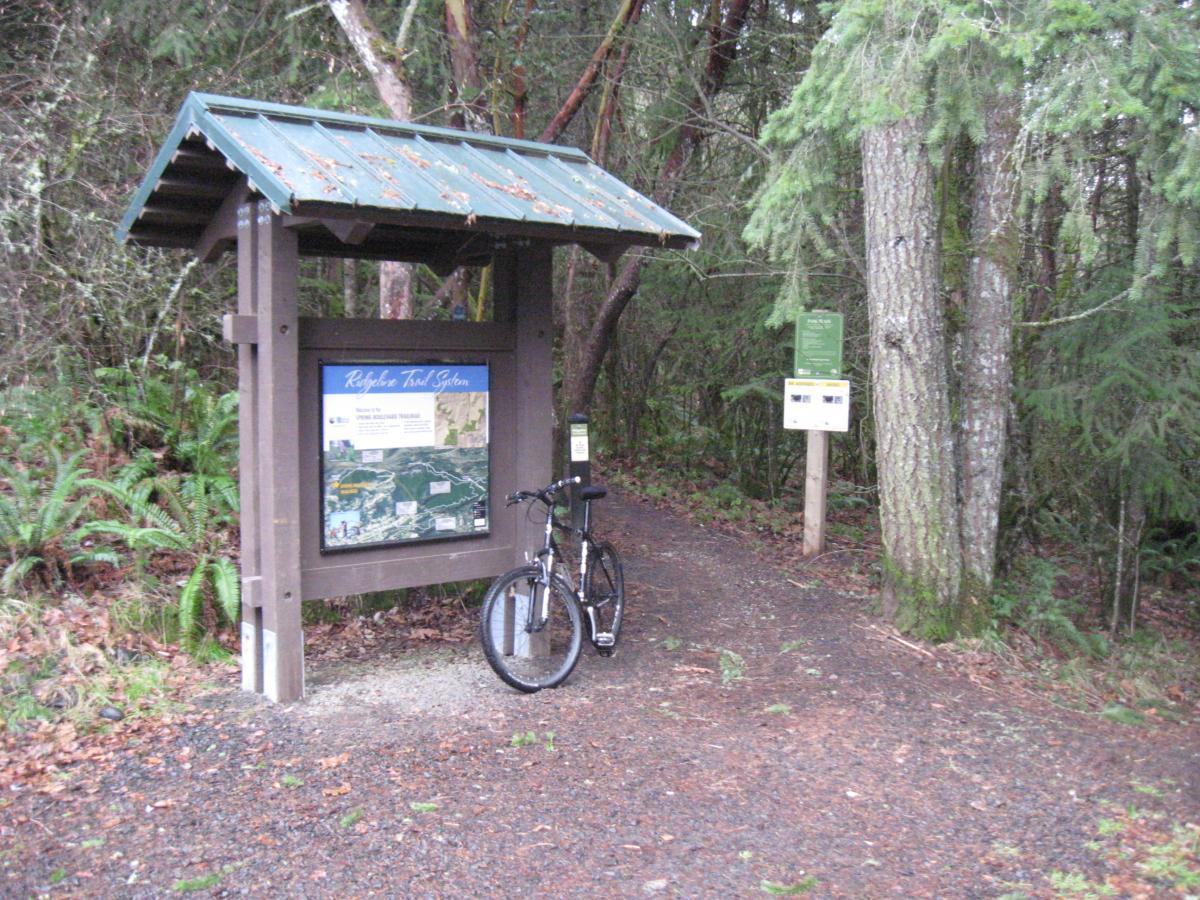 A trailhead sign for the Ridgetop Trail System, displaying a map of the trails. A mountain bike is resting against the sign, surrounded by lush greenery and trees. The path leads into a forested area, with additional signage nearby. Ridgeline Trail mountain bike trail.