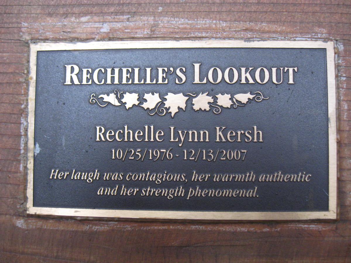 A bronze plaque celebrating Rechelle Lynn Kersh, inscribed with "Rechelle's Lookout." It includes her birth date (10/25/1976) and date of passing (12/13/2007), along with a heartfelt tribute: "Her laugh was contagious, her warmth authentic, and her strength phenomenal." The plaque is mounted on wood. Ridgeline Trail mountain bike trail.