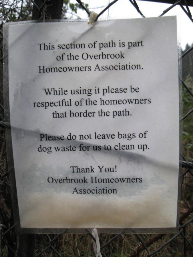 A printed sign attached to a fence, stating guidelines for a public path maintained by the Overbrook Homeowners Association. The sign politely requests users to respect the neighboring homeowners and reminds them not to leave bags of dog waste, expressing gratitude at the end. Ridgeline Trail mountain bike trail.
