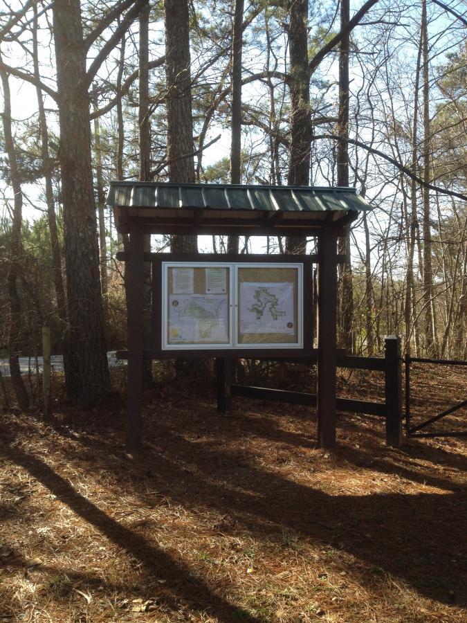 A wooden information sign with a green metal roof, displaying two maps, is situated in a wooded area with tall trees and pine needles on the ground. Sunlight filters through the branches, casting shadows on the ground. A fenced area is visible in the background, indicating access to a trail or natural site. Allatoona Creek Park mountain bike trail.