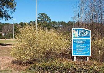 A blue and white sign for Lake Park, surrounded by green bushes, with a list of park amenities and information. In the background, there are trees and open grassy areas under a clear blue sky. Lake Rim Park Trail mountain bike trail.