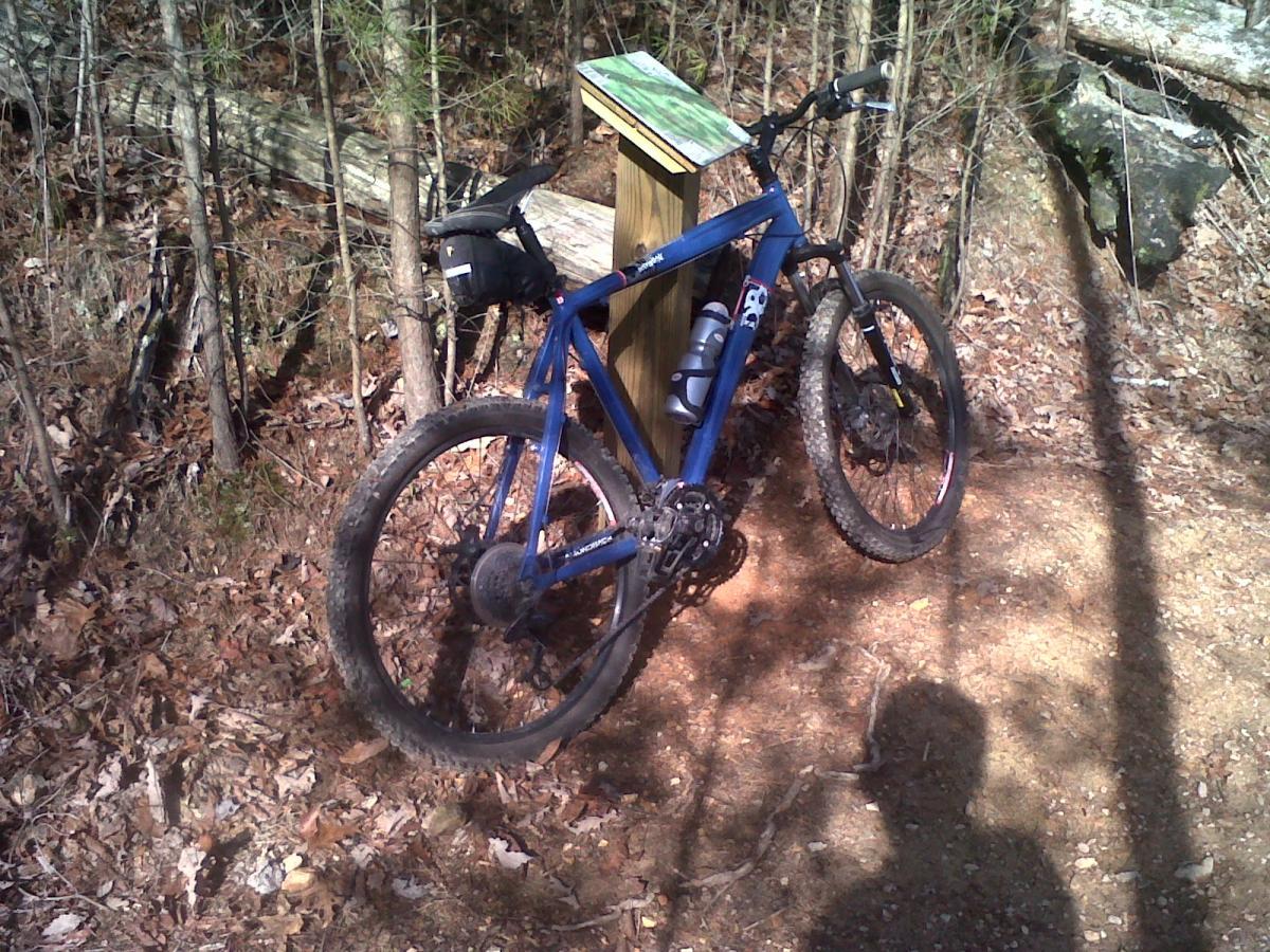 A blue mountain bike parked next to a wooden signpost in a wooded area, with fallen leaves and trees in the background. The bike has a water bottle mounted on it, and there is a shadow of a person visible on the ground. White Oak Mtn Biology Trails mountain bike trail.