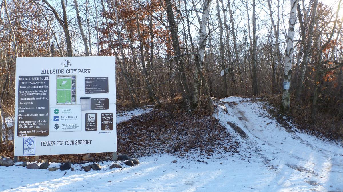 Signage at the entrance of Hillside City Park, displaying park rules, trail difficulty levels, and general information, with a snow-covered path leading into the woods. The sign includes sections for mountain biking, trail etiquette, and contact information for reporting issues. Surrounding trees are bare, indicating winter, with some leaves remaining on the branches. Hillside Park mountain bike trail.