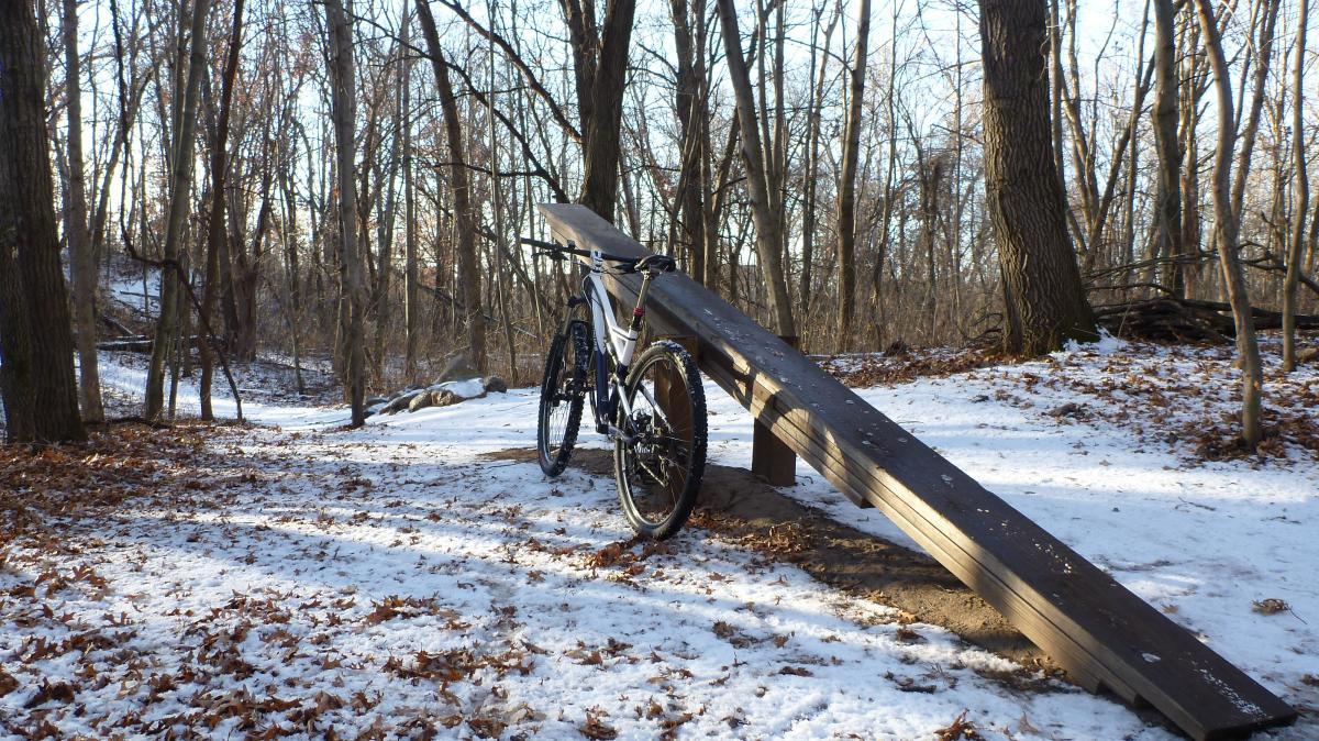 A mountain bike is resting next to a wooden ramp in a snowy forest setting, with scattered leaves and bare trees in the background. Hillside Park mountain bike trail.