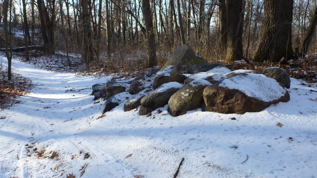 A snowy trail in a forest with a cluster of large, irregularly shaped rocks partially covered in snow. The surrounding trees are bare, indicating a winter landscape. The ground shows tire tracks and patches of fallen leaves along the path. Hillside Park mountain bike trail.