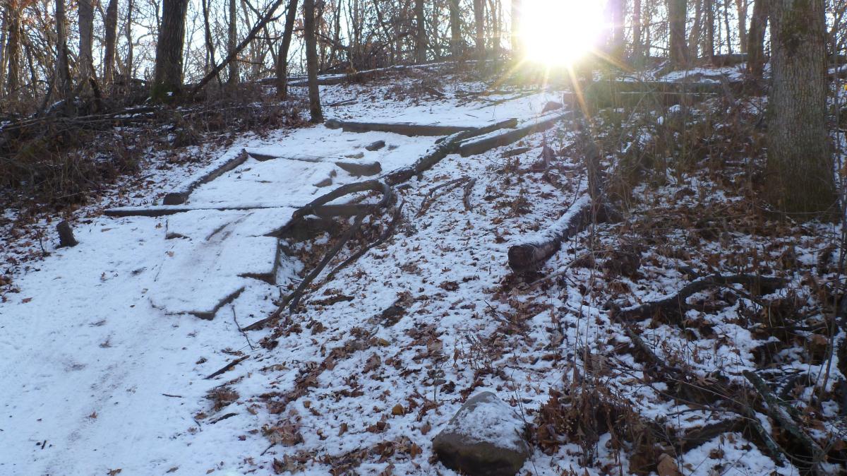 A snowy trail winding through a forest, with scattered fallen leaves and branches. The sun is shining through the trees, creating a bright light effect. The ground is partially covered in snow, with some exposed dirt and rocks visible along the path. Hillside Park mountain bike trail.