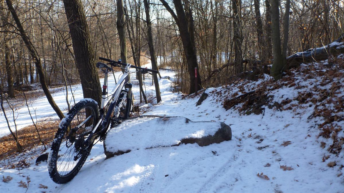 A mountain bike resting on a snow-covered rock along a winding trail in a wooded area, with bare trees and patches of snow on the ground. Hillside Park mountain bike trail.