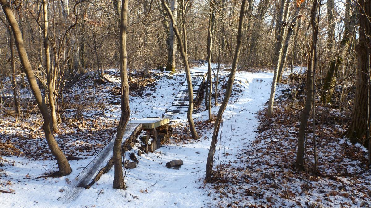 A snowy pathway winding through a forest, featuring a small wooden bridge over a gentle incline. The trees are bare, and fallen leaves cover the ground, creating a serene winter landscape. Hillside Park mountain bike trail.