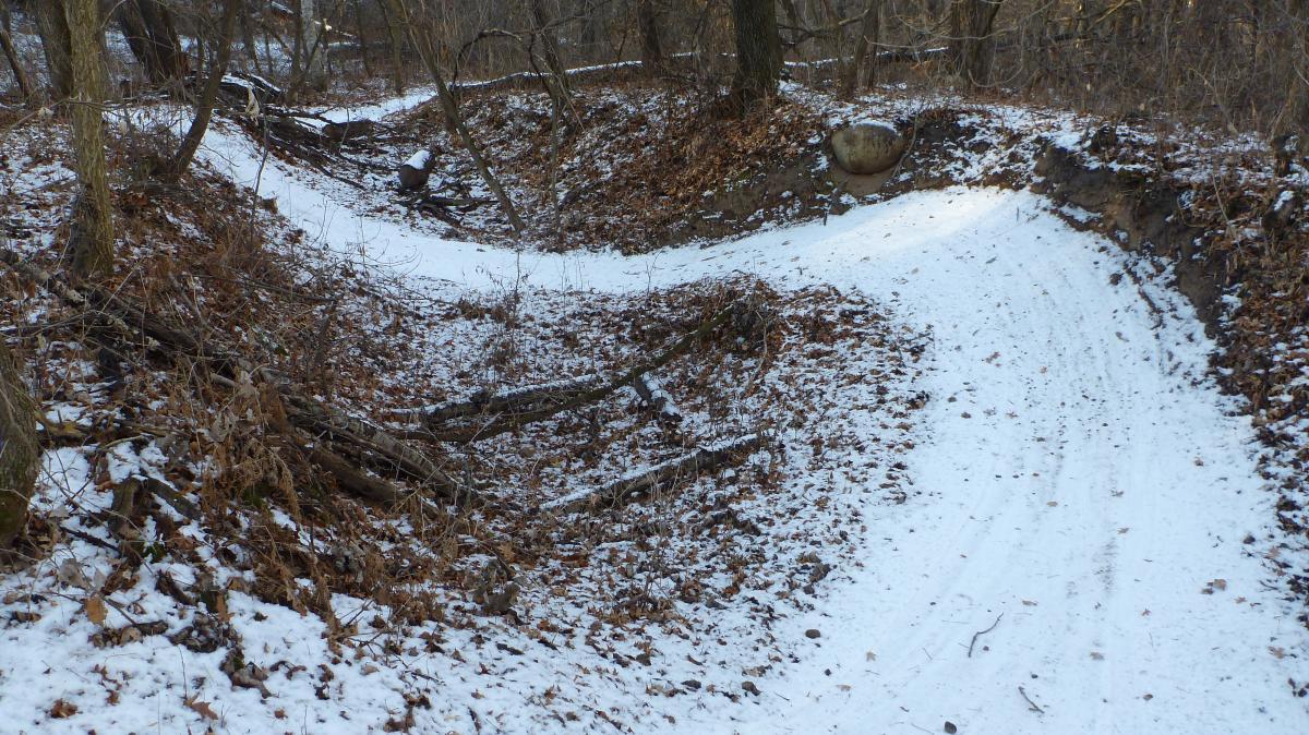A snowy forest trail curving through a wooded landscape, surrounded by brown leaves and fallen branches. The ground is partially covered in snow, and the terrain has steep banks on either side of the path. Hillside Park mountain bike trail.